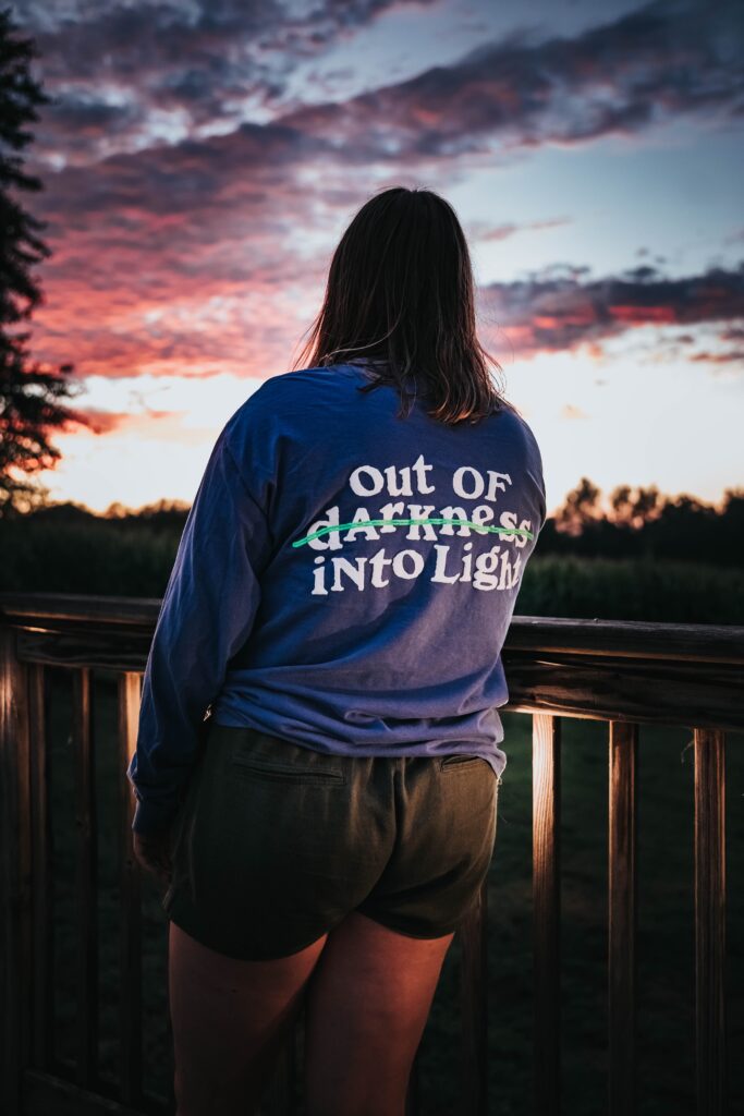 Woman in recovery looking out towards a sunset. Wearing a blue shirt that says “Out of Darkness into Light” with the word “darkness” scratched out so the shirt reads “Out of Darkness into Light.”